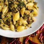 A white dish filled with shell pasta and fresh herbs sits on a wooden table, with red chili pieces scattered around for a warm, Kapha‑pacifying touch.
