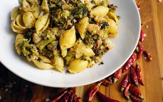 A white dish filled with shell pasta and fresh herbs sits on a wooden table, with red chili pieces scattered around for a warm, Kapha‑pacifying touch.