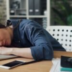 Tired woman resting her head on a laptop at an office desk, symbolizing workplace fatigue.